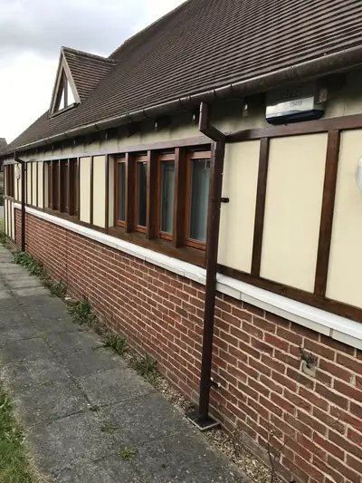 Long elevation of a building with dark stained timber framing, cream rendered panels, brick plinth and a stained downpipe.