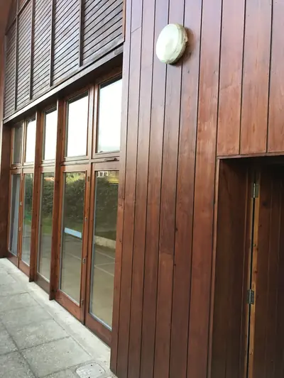 Corner of a timber-clad building finished in brown stain, with stained window frames and glazed doors along the elevation.