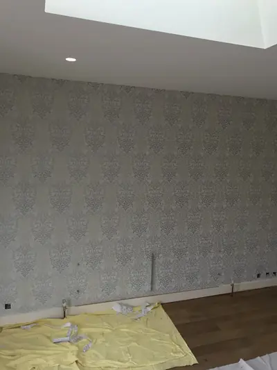 Newly hung silver damask wallpaper across a full wall, with sockets not yet refitted and a dust sheet and offcuts on the floor.