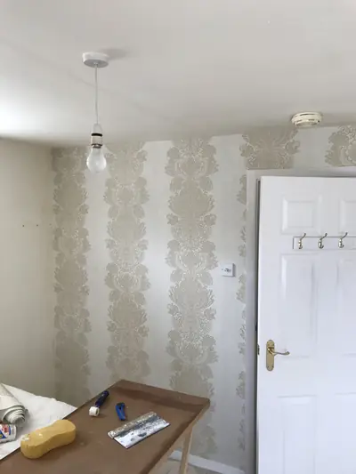 Bedroom being papered in a cream damask stripe, with a pasting table holding a smoothing brush, seam roller and sponge in front of it.