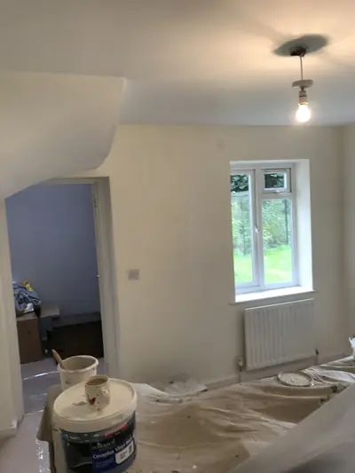 Cream painted room with a window and radiator, dust sheets across the floor and a tub of vinyl matt emulsion in the foreground.