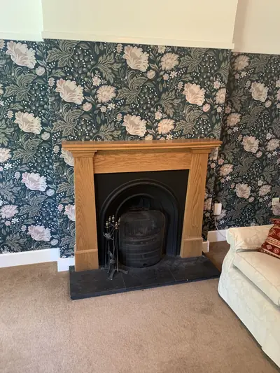 Dark green floral wallpaper covering a chimney breast and alcoves around an oak fire surround with a cast iron insert.