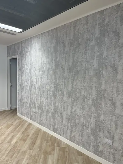 Office wall hung with silver-grey textured wallpaper running to a white painted skirting, with wood-effect flooring and a doorway at the left.