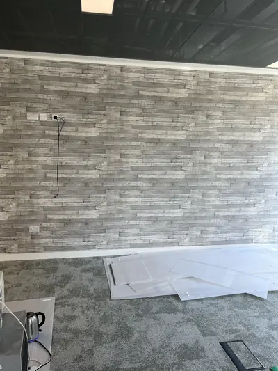 Office wall covered in grey weathered plank-effect wallpaper with sockets set into it, above a white skirting and grey carpet tiles.